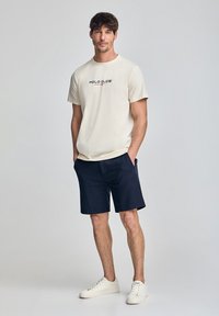 Kremowa koszulka z logo "POLO CLUB", zestawiona z granatowymi szortami i białymi trampkami, stojąca na tle jednolitym. Luźny fason, krótkie rękawy.