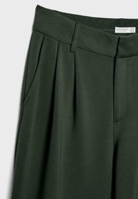 Pantalons vert foncé en tissu doux, dotés de plis, de poches latérales et d'une ceinture lisse avec une étiquette indiquant la marque et les informations de taille.