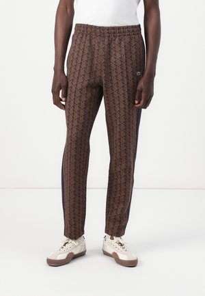 Brown patterned jogger pants with a geometric design, elastic waist, and navy side stripes. Paired with white sneakers.