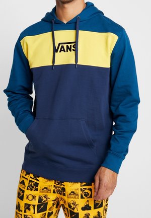 Homme portant un sweat à capuche Vans bleu et jaune avec des cordons de serrage, ainsi qu'un short jaune vif orné de carrés graphiques noirs.