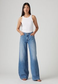 Top sin mangas blanco ajustado, jeans de pierna ancha azul de tiro alto con un acabado desteñido, que presentan bolsillos frontales y trabillas para cinturón, combinados con sandalias con tiras.