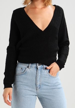 Femme portant un pull côtelé noir à col en V profond rentré dans un jean taille haute bleu clair, une main dans la poche, fond neutre.