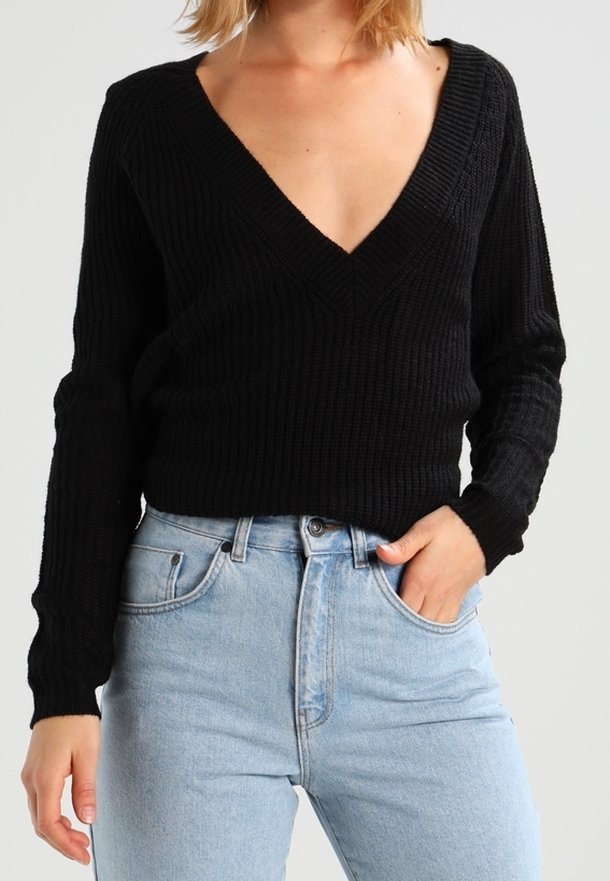 Femme portant un pull côtelé noir à col en V profond rentré dans un jean taille haute bleu clair, une main dans la poche, fond neutre.