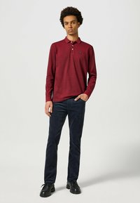 Bordeaux lange mouw polo met een kraag, een enkel borstlogo, gecombineerd met marineblauwe corduroy broek en zwarte veterschoenen, staande op een neutrale achtergrond.