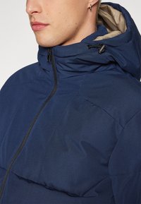 Giacca con cappuccio blu navy a finitura liscia e testurizzata, dotata di una zip frontale e cappuccio regolabile con cordino.