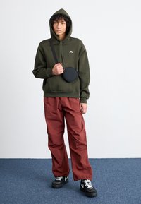 Green cotton hoodie with a front pocket, red lightweight pants, and a round black crossbody bag. Black shoes with white laces.