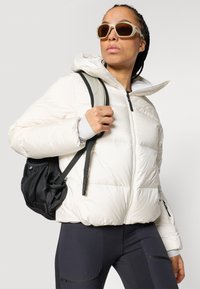 Krémová péřová bunda so zipsom vpredu, nastaviteľnou kapucňou a prešívanými detailmi. Čierny sieťovaný batoh a tmavé legíny dotvárajú vzhľad.