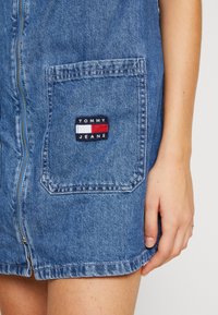 Dżinsowa spódnica z zamkiem z przodu i dużą kieszenią z logo Tommy Jeans, obok ręki i uda osoby.