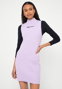 Moteris dėvi rankovių neturinčią alyvinės spalvos ribinę suknelę ant juodos ilgomis rankovėmis palaidinės, ant krūtinės yra užrašas „Karl Lagerfeld Jeans“.