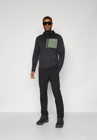 Schwarze Zip-Up-Jacke mit grüner Brusttasche, kombiniert mit schwarzen Hosen und robustem schwarzen Schuhwerk. Das Material scheint leicht und atmungsaktiv zu sein.