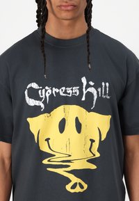 Čierne bavlnené tričko s veľkým žltým grafickým obrázkom usmievajúcej sa tváre s nerovnomerným, reflexným efektom a textom "Cypress Hill" v bielej farbe nad ním.