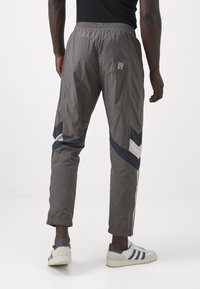 Joggers de nailon gris con rayas diagonales en azul marino y blanco, cinturilla elástica y puños en los tobillos. Presenta un bolsillo trasero con un logo.