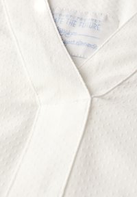 Tissu blanc avec un motif texturé, présentant un design en V et des coutures visibles. Une étiquette est cousue dans l'encolure.