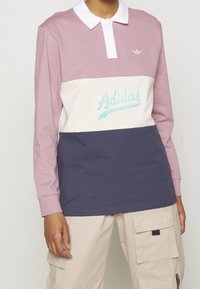 Person, der ein langärmliges, mehrfarbiges Adidas-Shirt in Mauve, Weiß und Marineblau trägt, kombiniert mit beigen Cargo-Hosen vor einem einfarbigen Hintergrund.