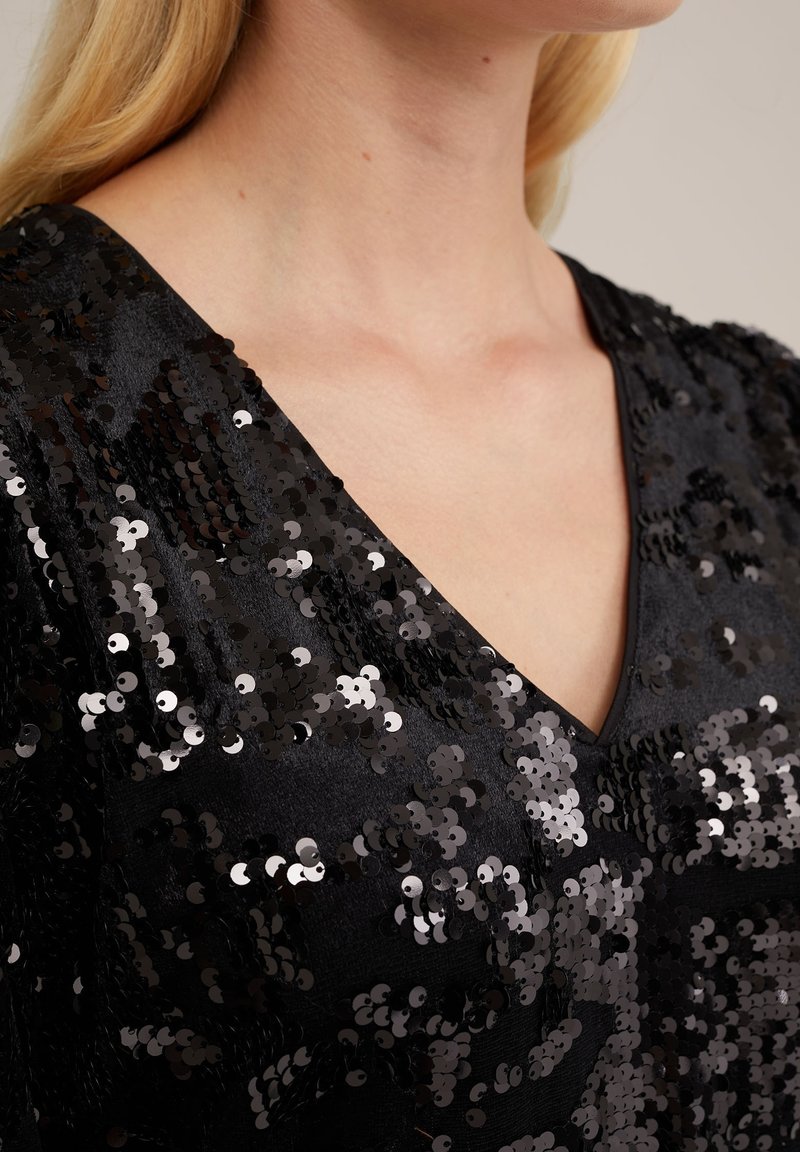 Schwarzes Pailletten-Oberteil mit V-Ausschnitt, aus einem strukturierten Stoff gefertigt, der mit glänzenden, überlappenden Pailletten versehen ist, die einen reflektierenden Effekt erzeugen.