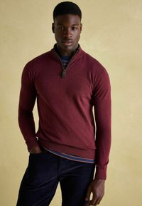 Burgundský sveter so zipsom a ribbovaným spodkom a manžetami, vrstvený cez námornícky pruhovaný košeľu, nosený s tmavými kordovým nohavicami.