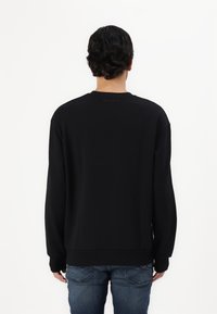 Crna dukserica s dugim rukavima, okruglim izrezom, rebrastim manžetama i ravnim donjim dijelom. Ima suptilan logotip brenda na leđima. U kombinaciji s plavim trapericama.