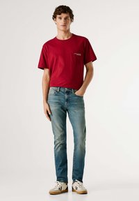 Rød bomulls-T-skjorte med lomme; lyseblå denimjeans; hvite joggesko med beige såle; krøllete hår; står mot en nøytral bakgrunn.