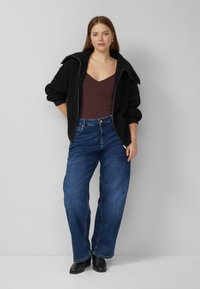 Zwarte, getextureerde jas met ritssluiting en brede revers, bordeauxrode V-hals top en losse blauwe spijkerbroek. Draagt zwarte laarzen, staand tegen een lichte achtergrond.