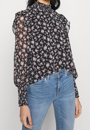 Vrouw draagt een zwarte doorschijnende blouse met lange mouwen, roze bloemenprint en ruches, gecombineerd met lichtblauwe jeans met hoge taille.