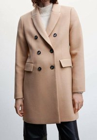 Abrigo beige de doble botonadura en tejido suave, con botones negros, solapas con muesca y bolsillos frontales. Textura lisa y corte entallado.