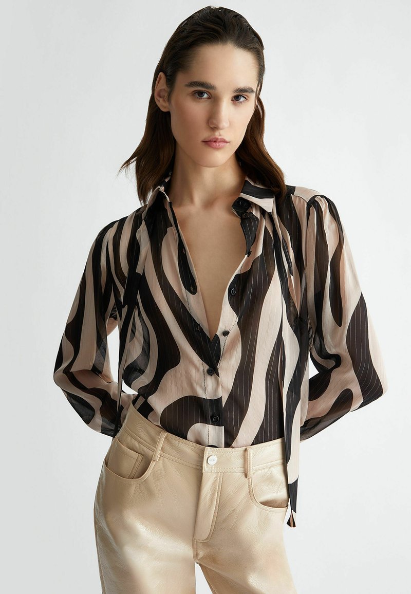 Blusa a maniche lunghe in tessuto trasparente con motivi a onde neri e crema, dotata di scollo a V profondo, patta con bottoni e maniche arricciate.