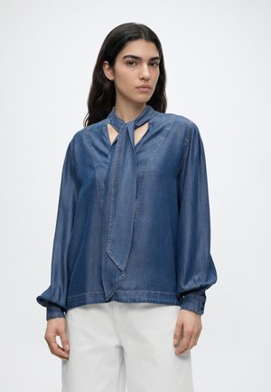 Donna con capelli lunghi e scuri che indossa una camicia di jeans blu con dettaglio annodato al collo e pantaloni bianchi, in piedi davanti a uno sfondo semplice.