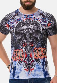 Grafisches T-Shirt mit komplexen schwarzen und roten Designs, Totenköpfen und Flügeln auf einem weißen und blauen Batik-Hintergrund. Kurze Ärmel, Rundhalsausschnitt.