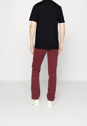 Personne debout de dos, portant une chemise noire à manches courtes, un pantalon bordeaux et des chaussures blanches sur un fond uni.
