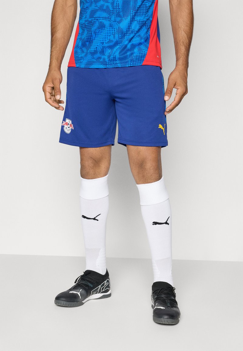 Puma sportsklær med en blå topp med grafisk mønster, røde detaljer, blå shorts, hvite sokker med svart logo og svarte sko.