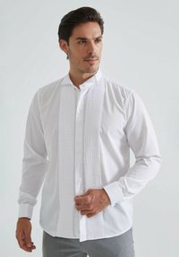 Chemise blanche à manches longues avec un devant plissé, un col classique et des poignets boutonnés. Tissu lisse avec une coupe décontractée ; sans motifs ni accents visibles.