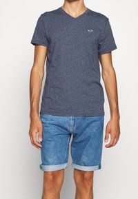 Námořnická modrá tričko s výstřihem do V vyrobené z texturované látky, spárované se světle modrými džínovými šortkami s ohrnutými rukávy a zelenými akcenty.