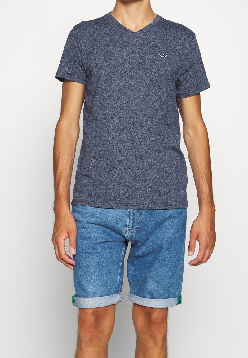 Námořnická modrá tričko s výstřihem do V vyrobené z texturované látky, spárované se světle modrými džínovými šortkami s ohrnutými rukávy a zelenými akcenty.