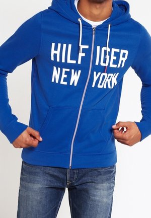 Man draagt een blauwe hoodie met rits en witte trekkoorden en de tekst "HILFIGER NEW YORK", gecombineerd met een blauwe spijkerbroek.