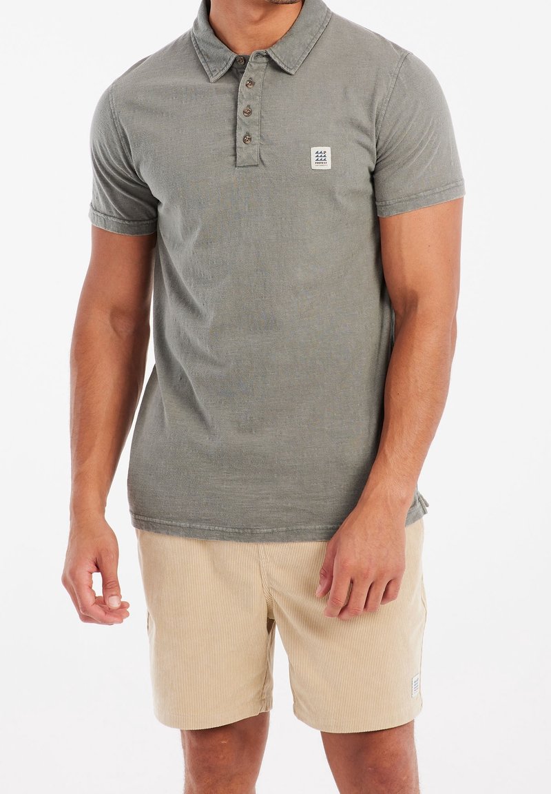 Homme portant un polo à manches courtes vert olive et un short en velours côtelé beige, debout devant un fond blanc.