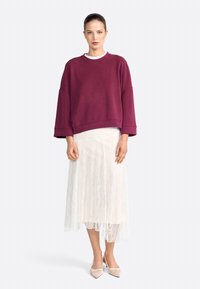 Felpa oversize bordeaux con spalle scese, abbinata a una gonna midi di pizzo bianca. Calzature: décolleté punta a cappio color crema.