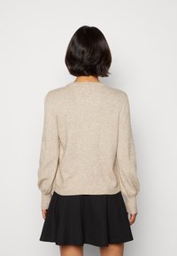 Pull en maille beige avec manches longues bouffantes et col rond, tissu texturé. Associé à une jupe évasée noire ; vu de dos.