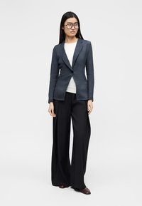 Žena s dugom crnom kosom i naočalama, odjevena u tamnoplavi blejzer, bijelu majicu, široke crne hlače i smeđe cipele, stoji.