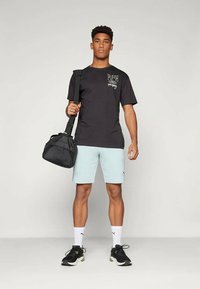 Černé grafické tričko, světlé modré kraťasy, nese černou sportovní tašku. Tenisky mají černé a zelené akcenty, bílé ponožky do kotníků. Jednoduchý, sportovní vzhled.