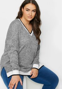 Maglione grigio a maglia con scollo a V, con bordi in nero e bianco sul collo e sui polsini. Indossato sopra pantaloni in denim blu.