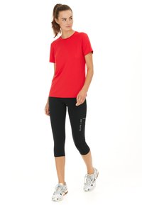 Vrouw staand, draagt een rood shirt met korte mouwen, zwarte cropped leggings met de tekst "ELITE LAB" en witte sportschoenen, kijkt opzij.