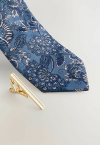 Cravate bleue à motifs floraux dans des teintes plus sombres, avec une pince à cravate dorée positionnée à proximité. Le tissu texturé ajoute de la profondeur.