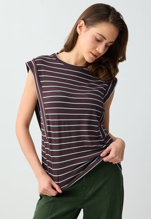 Femme portant un haut marron sans manches avec des rayures horizontales blanches, regardant vers le bas, tenant l'ourlet, associée à un pantalon vert sur fond clair.