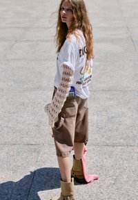 T-shirt blanc graphique avec impression colorée, associé à un short marron. Détail de manches longues en crochet et bottes beiges avec une accent rose.
