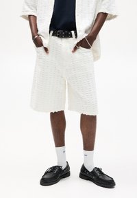 Pantaloni corti bianchi con una vestibilità ampia, abbinati a una cintura nera. Indossati con calzini bianchi e scarpe nere allacciare. Stile informale.