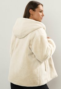 Ljust beige faux fur hoodie-jacka med avslappnad passform, som har en stor huva och sidofickor. Mjuk textur och inga synliga metalldetaljer.