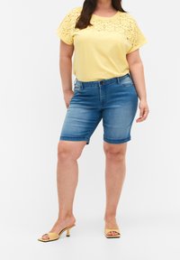 Žlutý krátký rukáv top s vyšívanými detaily, doplněný modrými denimovými šortkami a žlutými sandály na podpatku. Neformální letní outfit.