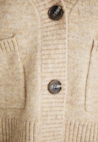 Μπεζ πλεκτό cardigan με ριγωτή υφή, μπροστινές τσέπες και δύο σκούρους καφέ κουμπιά. Μαλακό υλικό από μείγμα μάλλινων ινών.