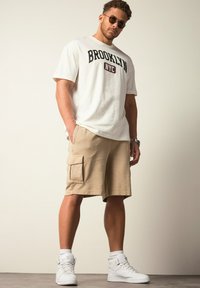T-shirt blanc avec le texte "BROOKLYN NYC", shorts cargo beiges avec poches latérales, baskets montantes blanches et lunettes de soleil, pose debout.