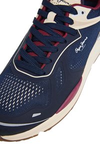 Mėlyna sportinė avalynė su akių tinklo viršumi, raudonomis detalėmis, plokščiais raišteliais ir gumine padu. Puošta firminiu logotipu ir tekstūruotomis intarpais, suteikiančiais atramą.